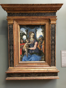 My image description: As the title describes, there are four figures in the painting who take up most of the lower half of the painting, with the Madonna located most centrally and prominently amongst them. She holds her baby, who stands somewhat upright on her right thigh and places his hands right above his mother’s nipple on her unclothed breast. The four figures have brightly-colored draped fabric garments while the landscape behind them is in ruins, with two large arches to either side of the Madonna that look like they are in a state of being of reclaimed by vegetation and forests. 