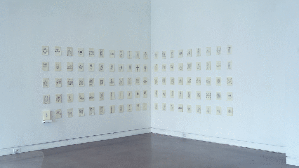 Wide-angle install photo of ninety-nine five by seven inch inch ink drawings of different museum object at the Metropolitan Art Museum drawn on handmade paper, along with one coloring book containing copies of each. Hung in a five row, twenty column grid at the corner of the white-walled, concrete floor gallery. The book sits on a small white ledge in the bottom left of the grid, accompanied by one red, yellow and blue colored pencils. 