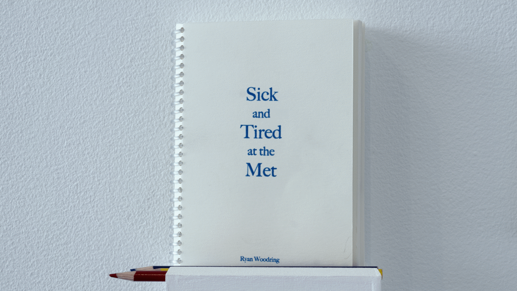 Detail photo of the five by seven inch, clear spiral bound "Sick and Tired at the Met" coloring book resting atop a small white ledge alongside red, blue, yellow colored pencils. Off-white cover with serif font spelling out the book's title and author. 