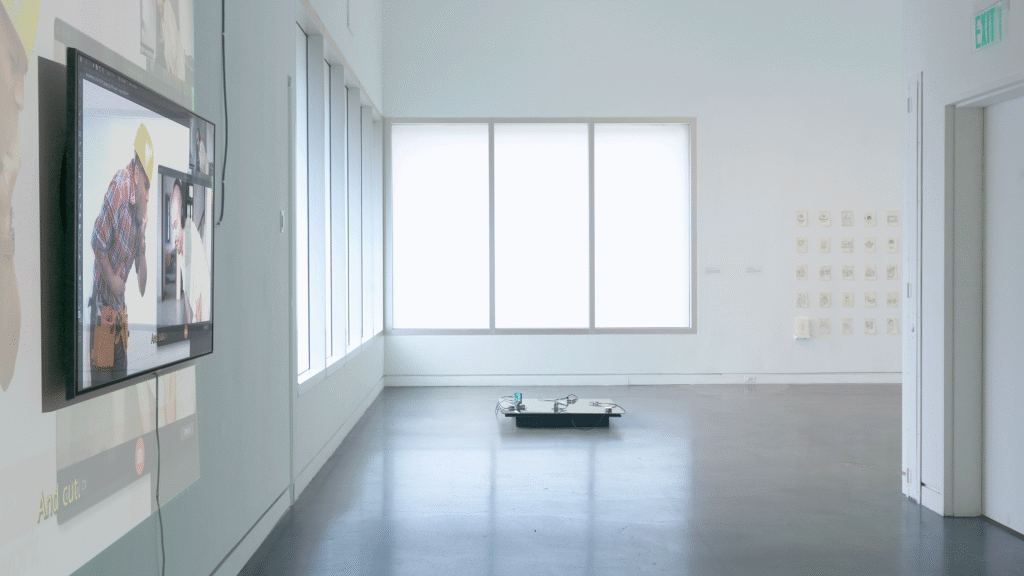 Wide-angle install photograph in a white-walled gallery with concrete floors and a band of tall frosted windows in the corner of the gallery letting in a cool white light. One floor-based work is in fully in frame, flanked by partially visible video and drawing installations. 