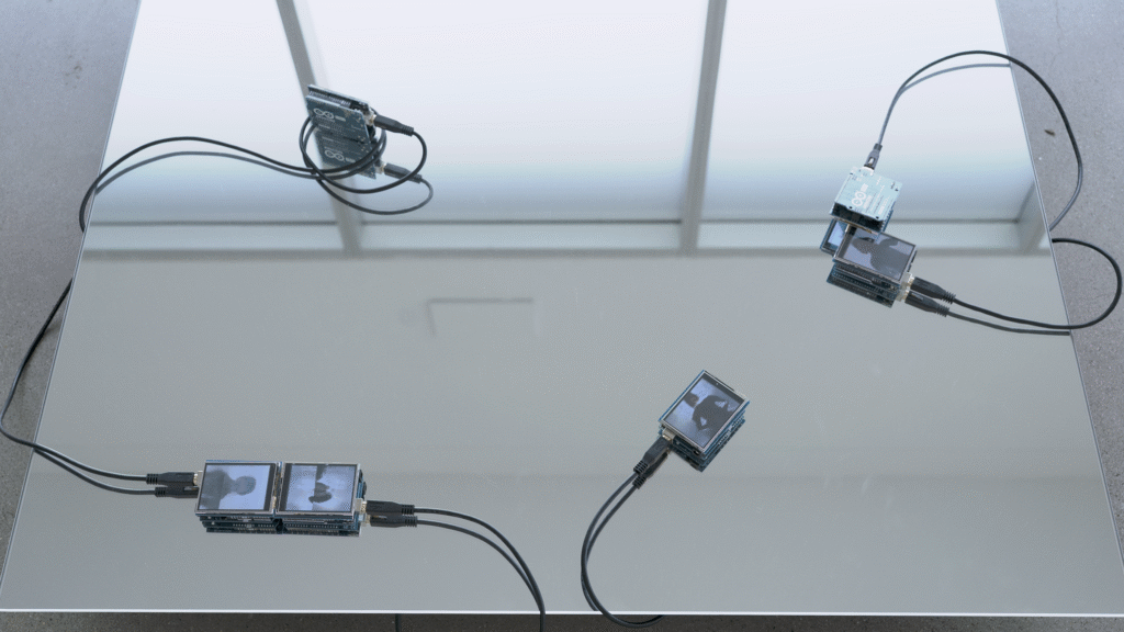 install detail photo shot from above a three by three foot mirror panel reflecting frosted windows and vaulted gallery ceiling. Resting atop the mirror are several small video displays connected to microprocessors and powered by snaking black usb cables that disappear under the mirror. Each video unit plays an image sequence of morphing portraits, with each subsequent frame arriving via a screen wipe.