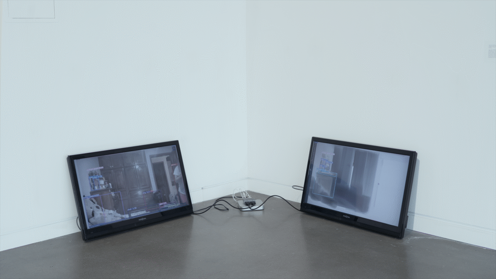 Medium distance install photo of a two-channel video installation in one corner of a white-wall, concrete floor gallery involving two resting forty inch monitors connected by a Mac mini and video splitter stationed between them. Each monitor leans on the wall behind it at a slight angle. The left screen displays a video feed of a bedroom with wood cabinets and bed in the foreground. The right screen displays an in-progress digital painting of the same scene.