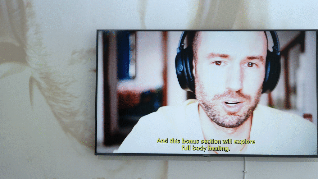 Detail install photo of a wall-mounted sixty-inch monitor. A still from the short film depicts the artist with headphones and yellow caption text: "And this bonus section will explore full body healing." The same image is faintly projected at a larger scale onto the same wall as the monitor. 