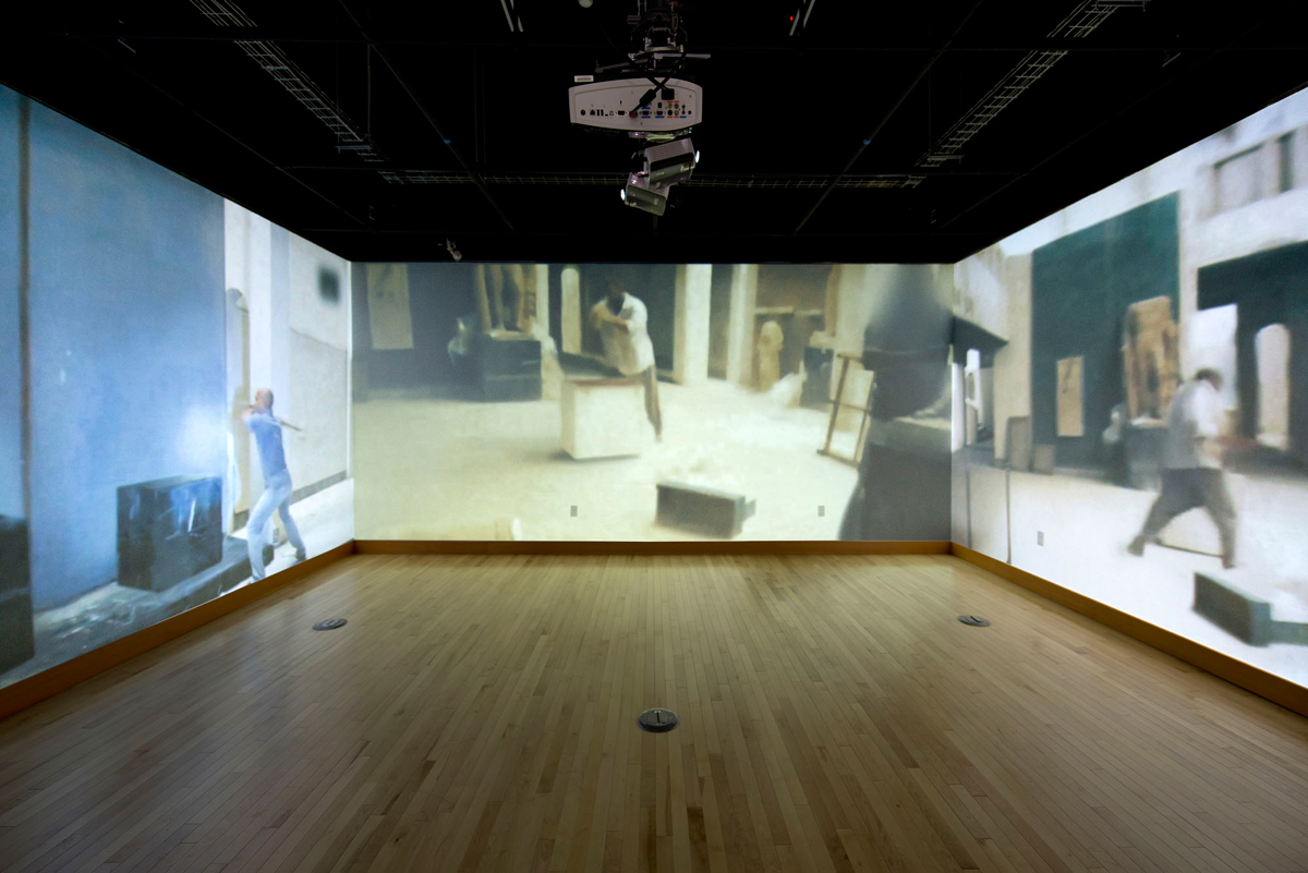 a centered, wide angle photo showing 3 walls of a 4 wall video installation at White Box Gallery in Portland, Oregon. The four walls each show one unique clip from an ISIS propaganda video of the destruction of artifacts at the Mosul Museum in Iraq, except in this version the scenes have, through visual effects manipulation, been emptied of the targeted artifacts.