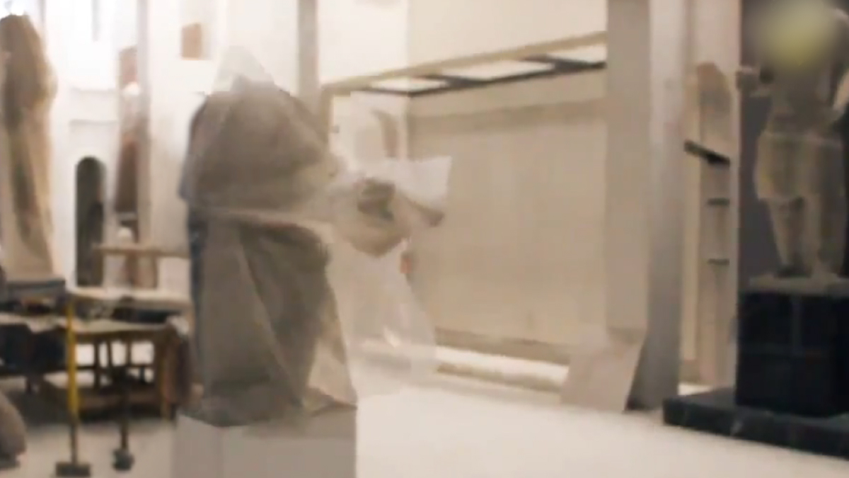 A still from a video clip showing a sculpture of Venus, partially occluded by the thick plastic covering draped on it. The video is a 5 second loop of a scene from an ISIS propaganda video showing the destruction of artifacts at the Mosul Museum. In this version, the human perpetrators have been removed. The statue appears to be magically throwing off its own semi-transparent protective material to reveal itself.