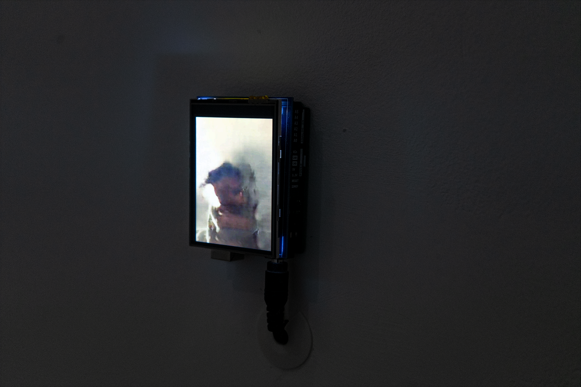 A closeup photo of a bright LCD displaying a static image that loosely resembles a person. The screen is attached to a microcontroller and hangs on the wall with four pushpins. A 12 volt power supply connects at the bottom of the microcontroller and then threads into the wall.