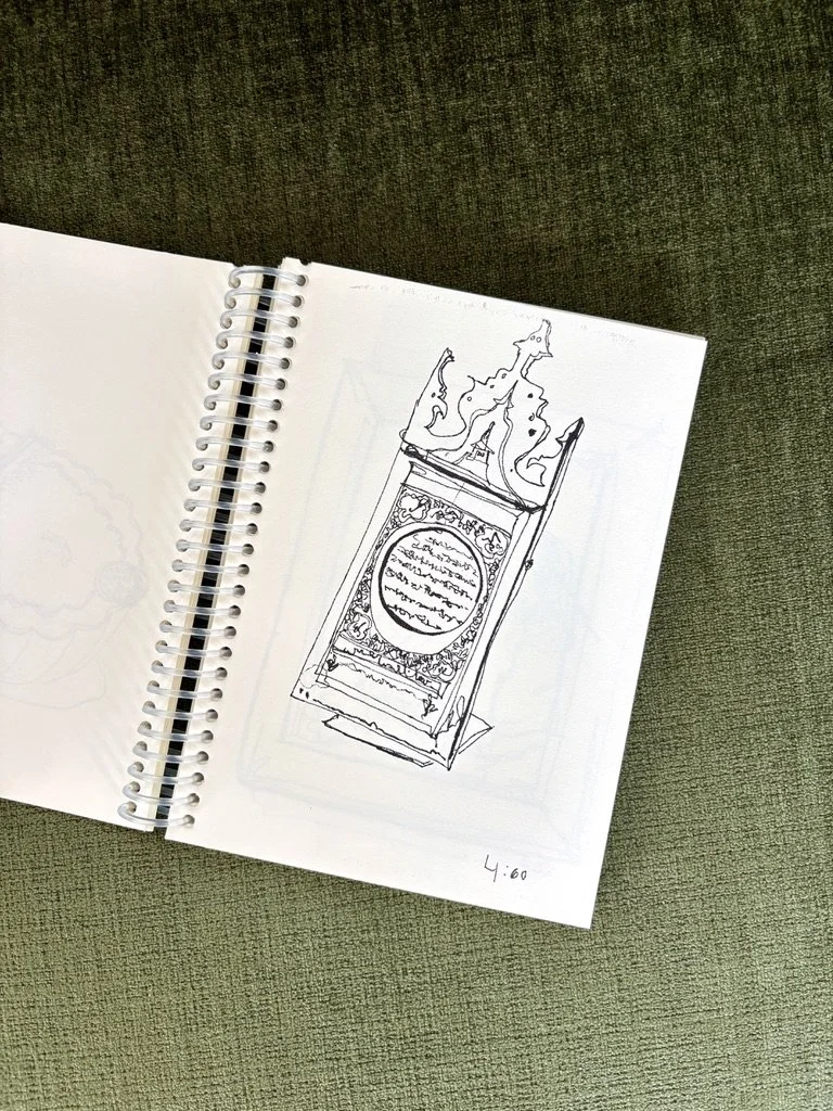 Coloring book lays open on a velvety green surface. Black ink contour line drawing of an ornately decorated, vertically-oriented placard on a stand. In the middle, a framed out circle filled with text-like squiggles.