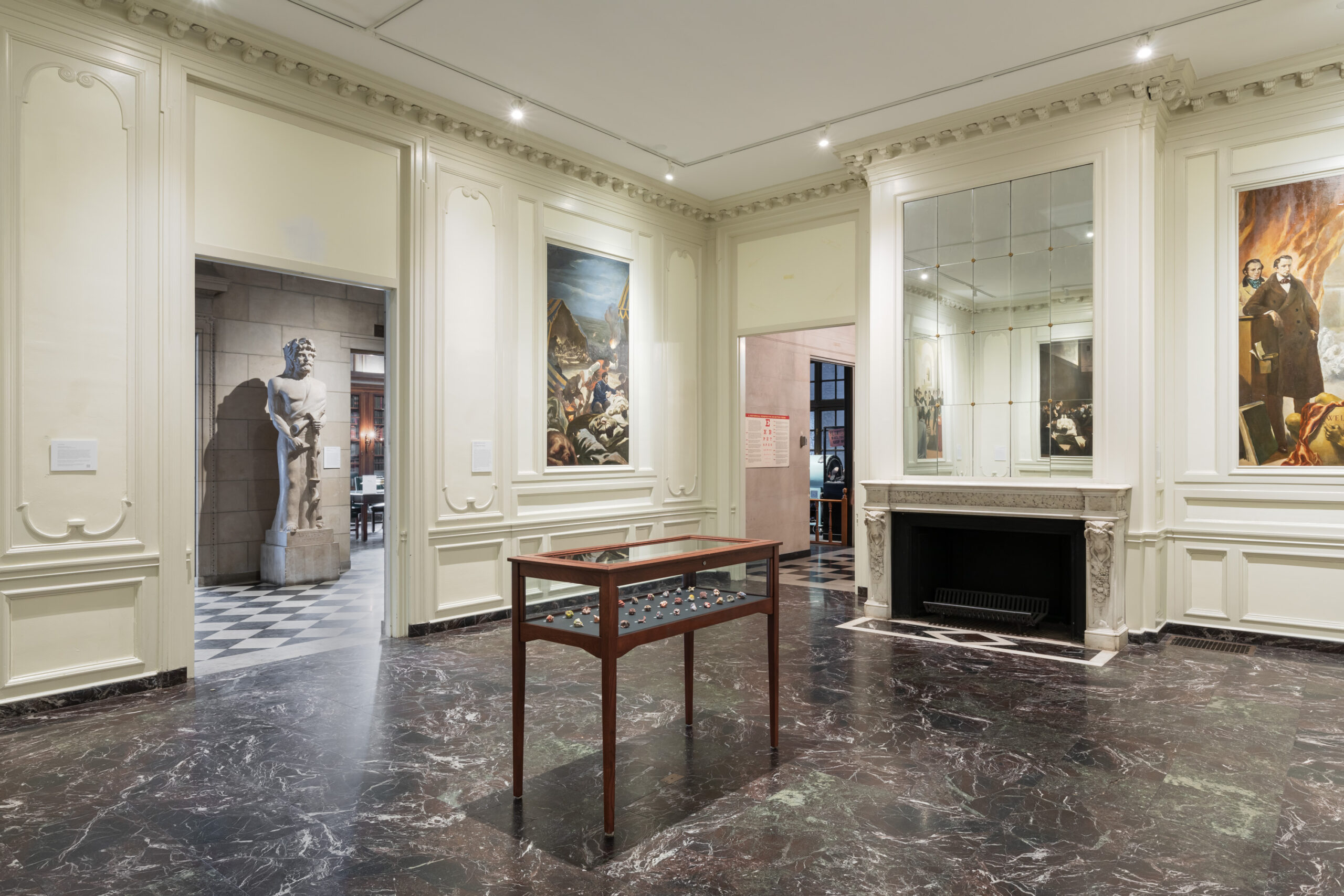 a wide angle photo taken from one corner of a large off-white room with ornate molding and vertically-oriented murals displaying different scenes from medical history like autopsies, deliveries, and academies. 2 murals are visible on the walls and another 2 in a large mural above a closed fireplace. A sleek wooden display case with clear glass panels rests in the center of the grand room atop a black, white and green marble stone floor. Inside the display case rests 30 small 3d-printed candies in a variety of colors. The room has two large doorframes that lead to other hallways in rooms in the museum.
