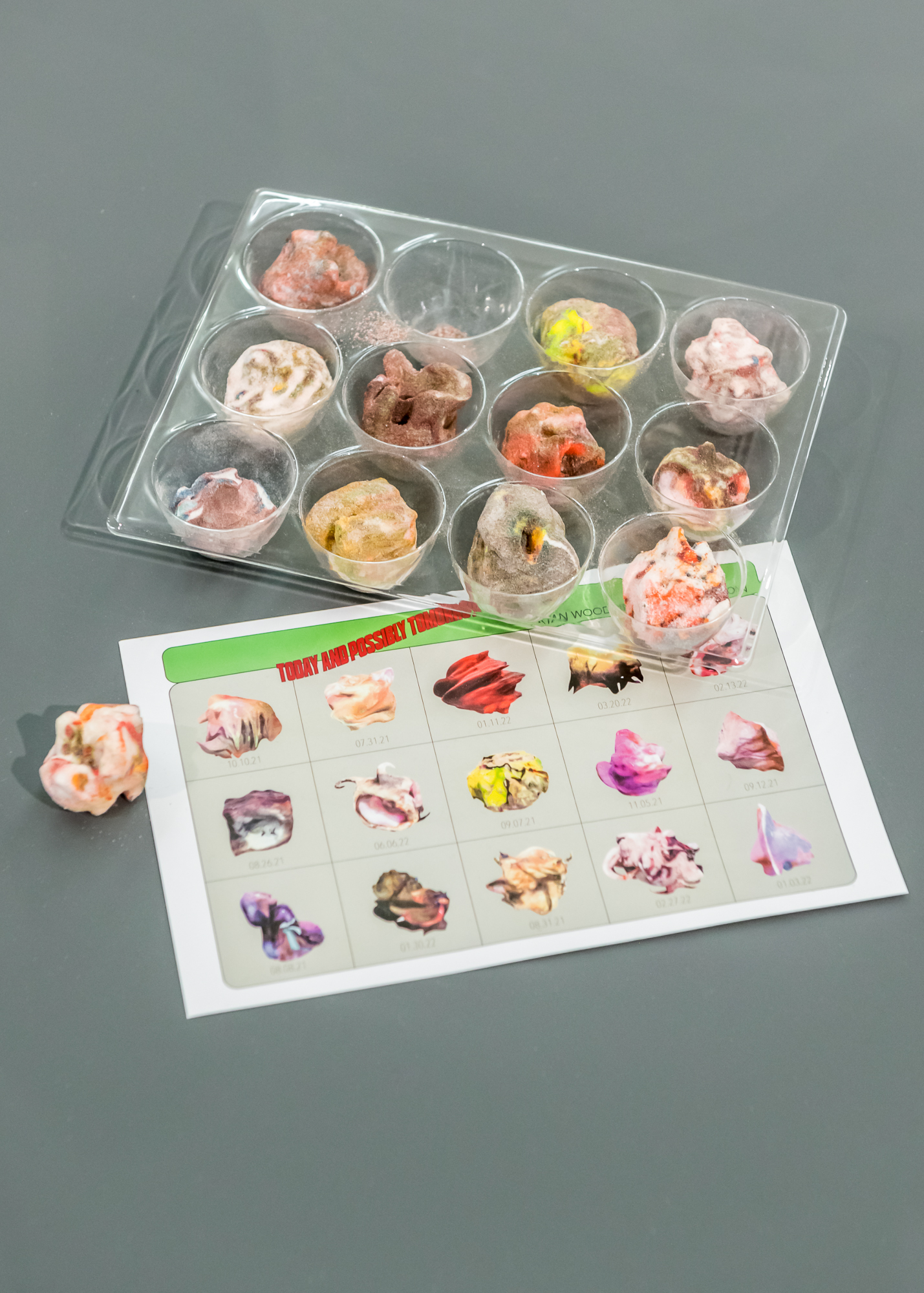 Vertically oriented overhead installation photograph at Sfaffordshire showing a plastic tray holding 11 3D-printed hard candies. Each is roughly the same volume while different in color and form. Sitting below the tray is an escaped candy and a printout detailing 30 different varieties of the candy in graphic form, each of them with the date of its inception beneath it.