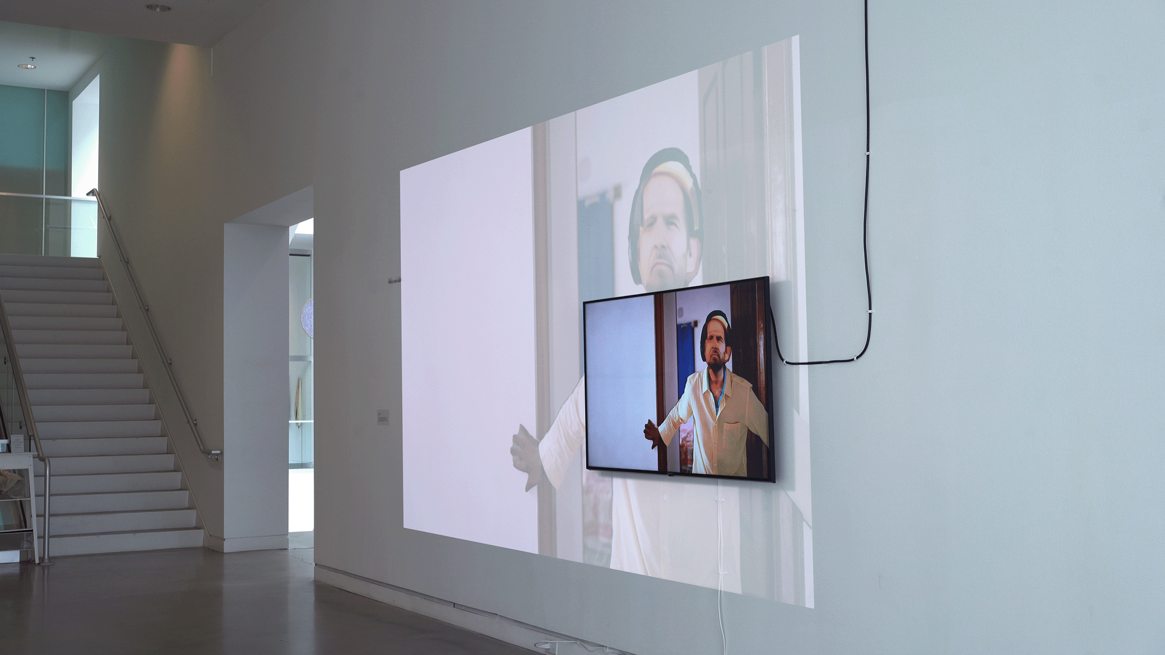 Wide-angle documentation of a two-channel video installation along a large white wall in a museum corridor. The installation includes a sixty inch wall-mounted monitor displaying a climactic handheld scene in which the camera follows a flickering-faced man as he slowly backpedals towards, and ultimately collapses on a bed. The monitor feed is echoed by a larger, fainter projection of the same video along the same wall.