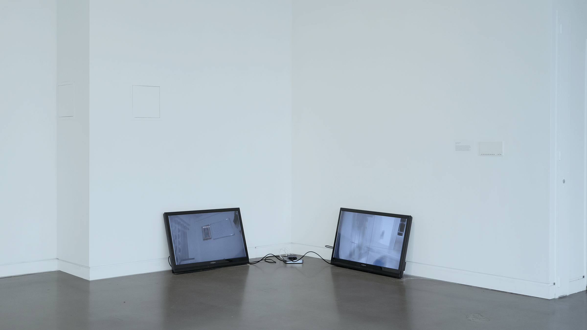 Wide-angle documentation of a synced two-channel video installation in one corner of a white-wall, concrete floor gallery. Two floor-resting forty inch monitors lean on their respective catty-corner walls, connected by a Mac mini, video splitter and HDMI cables stationed between them. One screen (left) displays a video feed from an online study site of unoccupied bedroom and offices. The other screen displays in-progress digital paintings of those scenes.