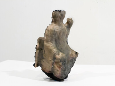 a medium close up image of a full color 3D print of a seated figure atop a white pedestal and white wall. The figure, barely recognizable as such due to the aging of the original sculpture and the lossy process of photogrammetry, appears draped in fabric and staring forward. The 3D print appears incomplete, with only one half in color and the other a jagged black color.
