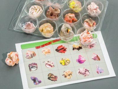 Vertically oriented overhead installation photograph at Sfaffordshire showing a plastic tray holding 11 3D-printed hard candies. Each is roughly the same volume while different in color and form. Sitting below the tray is an escaped candy and a printout detailing 30 different varieties of the candy in graphic form, each of them with the date of its inception beneath it.