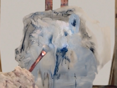 A medium close-up still from a video shot using a built-in laptop camera. A chunk of ice rests on an easel in the middle of the screen. From screen left a polar bear arm holds a paintbrush in front of the block of ice. A representational image of a polar bear appears on the ice.