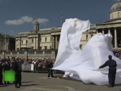 Still from a video of Trafalgar Square, London. A wide angle shot recorded from a tripod-mounted camera. A number of figures are pulling down a silky white fabric, which appears to have been cloaking something.