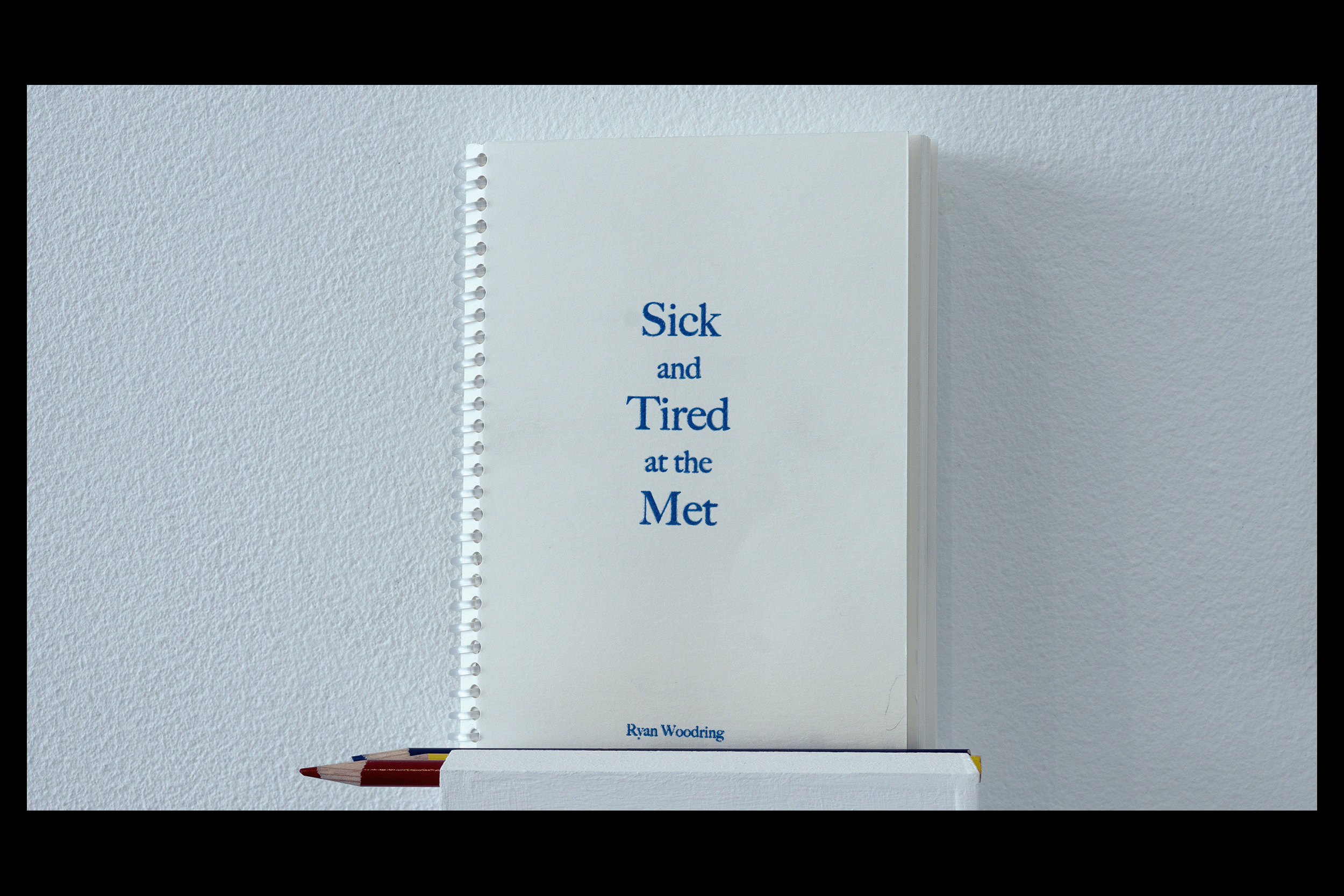 Detail photo of the five by seven inch, clear spiral bound 'Sick and Tired at the Met' coloring book resting atop a small white ledge alongside red, blue, yellow colored pencils. Off-white cover with serif font spelling out the book's title and author.