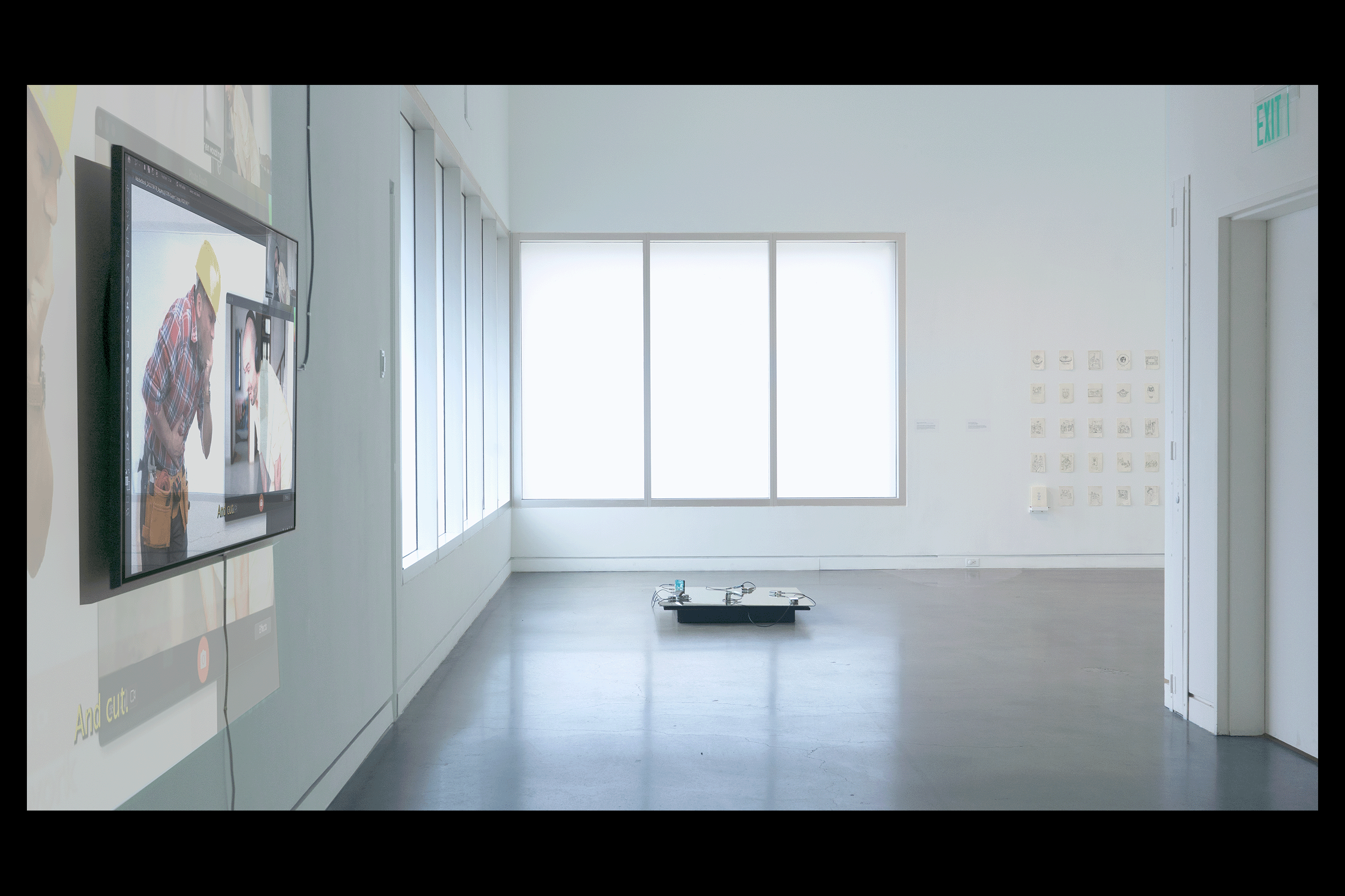 Wide-angle install photograph in a white-walled gallery with concrete floors and a band of tall frosted windows in the corner of the gallery letting in a cool white light. One floor-based work is in fully in frame, flanked by partially visible video and drawing installations.