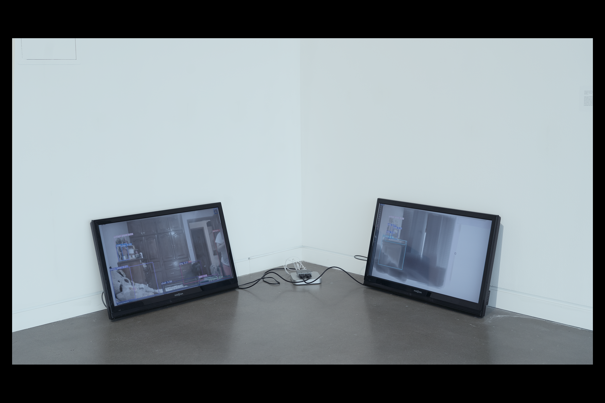 Medium distance install photo of a two-channel video installation in one corner of a white-wall, concrete floor gallery involving two resting forty inch monitors connected by a Mac mini and video splitter stationed between them. Each monitor leans on the wall behind it at a slight angle. The left screen displays a video feed of a bedroom with wood cabinets and bed in the foreground. The right screen displays an in-progress digital painting of the same scene.