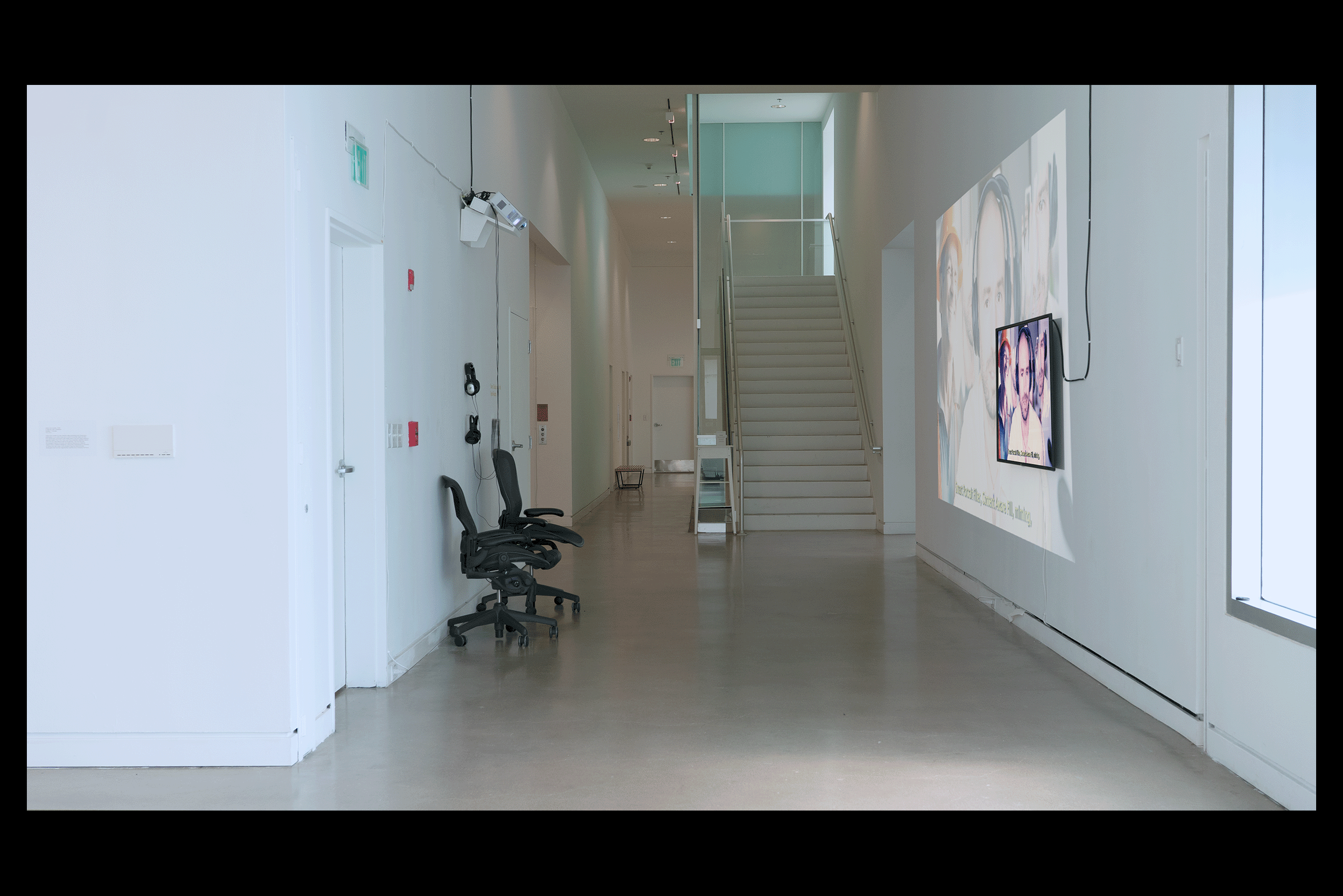 A wide angle install photo of a two-channel video installation along a long corridor wall with white walls and a concrete floor. Devoid of ceiling light, a cool ambient light from frosted windows sweeps through the corridor. A video still of the artist from the short film is displayed on a wall-mounted sixty inch monitor. The same image is faintly projected at a larger scale onto the wall behind the monitor. A black wire extends outwards and up from the monitor. On the other side of hallway are two black computer chairs and two pairs of black studio headphones. A staircase drenched in turquoise and orange hues is visible towards the center background.