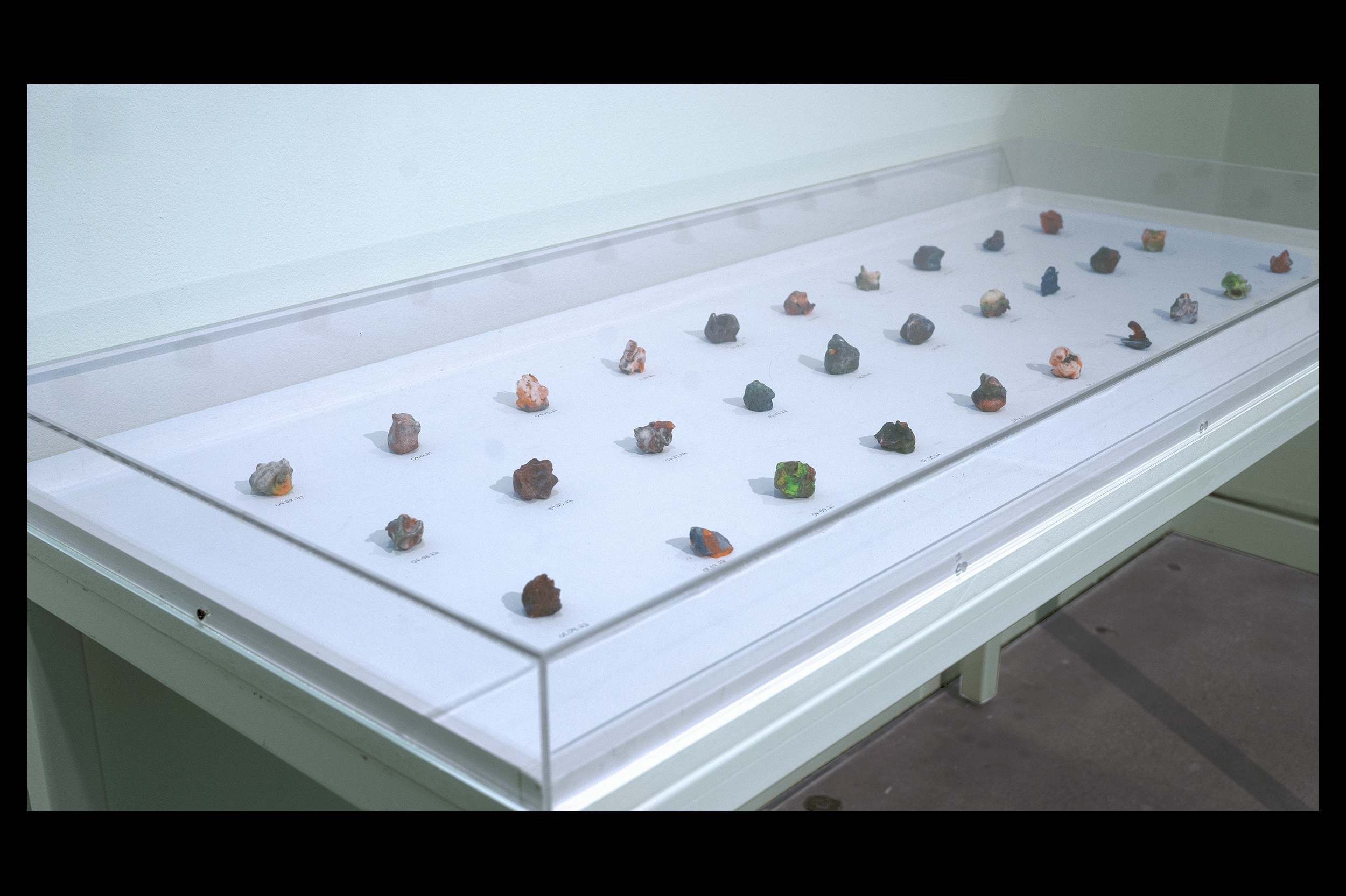 Install detail photo taken from above the front left corner of the clear plexi museum display case housing thirty 3D-printed hard candies that sit atop a white wooden surface accompanied by individual hand-written dates marking the date of each candy's modeling. The candies, greatly varying in shape and color, are roughly the volume of large gumballs and the consistency of condensed sugar.